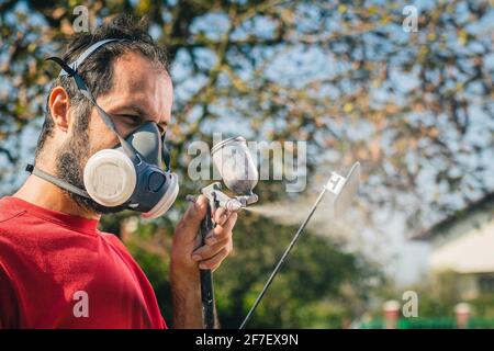 Pittore dilettante in maglia rossa e maschera facciale con una pistola a spruzzo per dipingere un oggetto di plastica o una parte in un giardino domestico. Profilo di un pittore con un mq Foto Stock