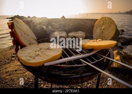 Grigliare hamburger su una piccola griglia in metallo portatile a carbone sulle rive del Lac Leman in Svizzera durante la prima sera d'inverno con il sole tramontare. Foto Stock