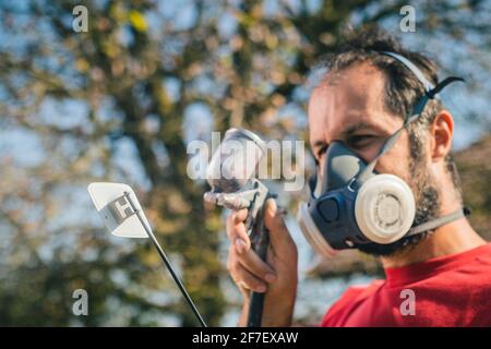 Pittore dilettante in maglia rossa e maschera facciale con una pistola a spruzzo per dipingere un oggetto di plastica o una parte in un giardino domestico. Profilo di un pittore con un mq Foto Stock