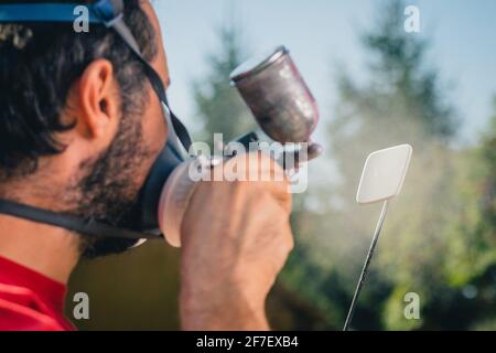 Pittore dilettante in maglia rossa e maschera facciale con una pistola a spruzzo per dipingere un oggetto di plastica o una parte in un giardino domestico. Vista posteriore di un pittore con un piccolo Foto Stock