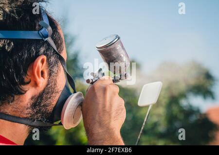 Pittore dilettante in maglia rossa e maschera facciale con una pistola a spruzzo per dipingere un oggetto di plastica o una parte in un giardino domestico. Vista posteriore di un pittore con un piccolo Foto Stock