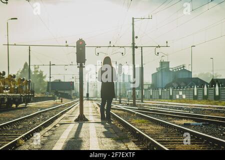 La giovane donna in giacca attende un treno su una stretta piattaforma di stazione tra le piste nelle prime ore del mattino subito dopo l'alba, con nebbia e malessere Foto Stock