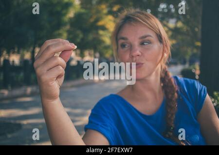 Una giovane donna caucasica che tiene una noce e la guarda mentre si gode il sole in un parco a Budapest, Ungheria mentre indossa una camicia blu. Foto Stock