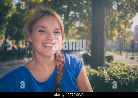 Una giovane donna caucasica sorridente e godente il sole d'autunno in un parco a Budapest, Ungheria mentre indossa una camicia blu. Foto Stock