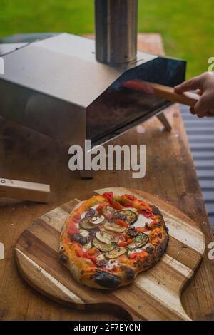 Deliziosa pizza fresca fatta in casa su un piatto rotondo di legno, direttamente da un forno portble domestico di acciaio inossidabile alimentato dai pellet. Pizza party all'aperto. Foto Stock