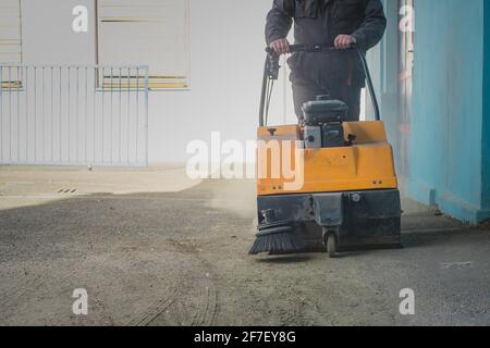 Una persona sta spingendo una spazzatrice industriale alimentata a benzina arancione su asfalto sporco nel tentativo di pulirlo da detriti e sabbia Foto Stock