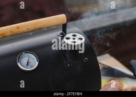 Fumo che esce da un fumetto di un piccolo fumatore nero griglia o barbecue, visibile anche indicatore di temperatura e maniglia di apertura in legno. Foto Stock