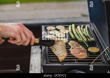 Uomo che gira carne di pollo con tenaglie in un piccolo fumatore nero griglia o barbecue, caricato con carne di pollo e verdure. Foto Stock