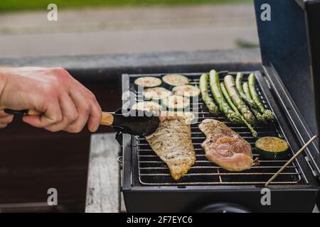 Uomo che gira carne di pollo con tenaglie in un piccolo fumatore nero griglia o barbecue, caricato con carne di pollo e verdure. Foto Stock