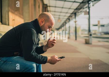 Vista laterale di un giovane maschio calvo seduto su una panchina e utilizzando un telefono e fumare mentre si attende un treno su una stazione ferroviaria vintage di stile più vecchio. Foto Stock