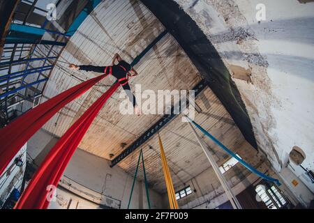 Una danzatrice da ginnastica aerea femminile su corda di seta che si esibisce in un ambiente indistrale di un laboratorio o magazzino abbandonato. Foto Stock