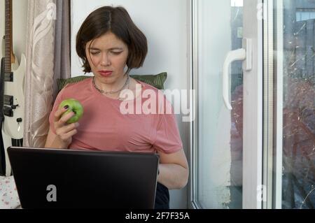 Donna attraente che lavora a distanza: Siediti sul davanzale con il laptop e mangia la mela. Foto Stock