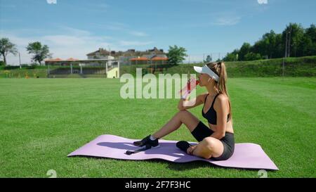 Vista laterale della giovane atletica donna che beve acqua dopo l'allenamento sul tappetino, campo stadio in estate soleggiato giorno. Fit ragazza che indossa abbigliamento sportivo che riposa, gustando una bevanda fresca. Concetto di sport, allenamento. Foto Stock