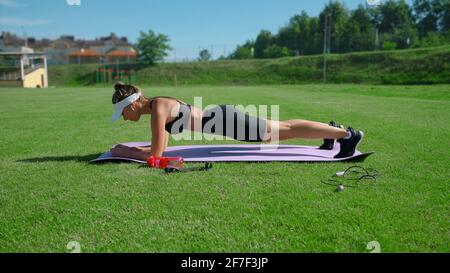 Vista laterale di una giovane donna concentrata che indossa abiti sportivi, berretto e cuffie wireless mantenendo la posizione della tavola. Ragazza di allenamento muscoli addominali, esercizio all'aperto su campo stadio con erba. Foto Stock