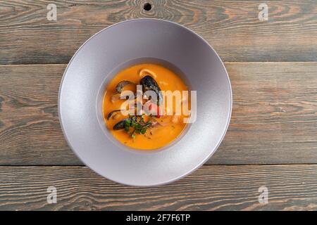 Dall'alto si può ammirare una gustosa zuppa d'arancia luminosa con cozze in conchiglie nere in piatto bianco su tavolo da ristorante in legno. Deliziosi frutti di mare serviti con germogli e pomodori. Concetto di esotica cucina asiatica. Foto Stock