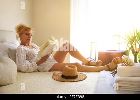 Donna felice in vacanza rilassata e sdraiata su un letto in una camera d'albergo che legge un libro in mano Foto Stock
