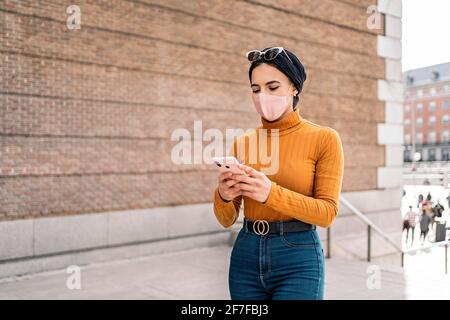 Bella giovane donna musulmana indossando sciarpa testa e maschera protettiva viso utilizzando il suo telefono cellulare in strada. Foto Stock