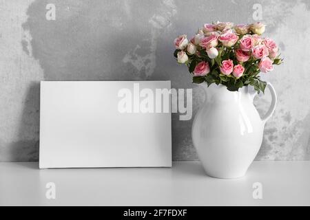 Mockup in tela bianca su tavolo bianco con fiori rosa in vaso su fondo parete in cemento. Tela stirata bianca, arredamento interno Foto Stock