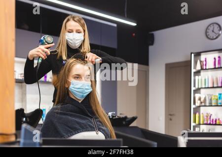 Master donna parrucchiere in una maschera medica asciuga i capelli della ragazza con un asciugacapelli e pettini dopo il lavaggio in un salone di bellezza. Covid-19 pandemia e. Foto Stock