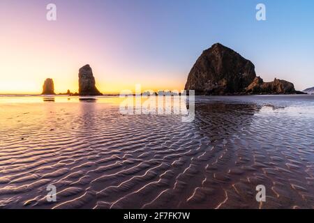 Haystack Rock e gli aghi al tramonto, con sabbia testurizzata in primo piano, Cannon Beach, contea di Clatsop, Oregon, Stati Uniti Foto Stock