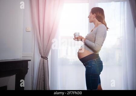 Donna in piedi vicino alla finestra, bere tè guardando fuori Foto Stock