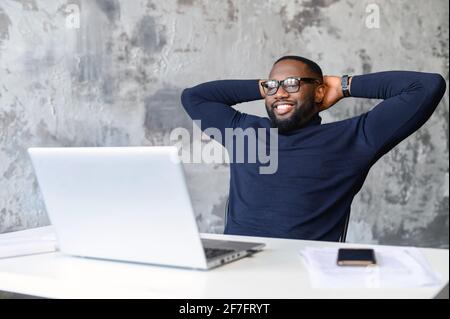 Un allegro dipendente dell'ufficio afroamericano di sesso maschile si trova sul posto di lavoro e si è appoggiato di fronte al computer portatile. Un freelance soddisfatto e dalla pelle scura ha fatto il progetto e si rallegra Foto Stock