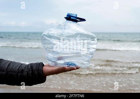 primo piano di un giovane uomo caucasico di fronte al ocean tenendo una bottiglia di plastica usata in mano di fresco raccolto dalla sabbia di una spiaggia Foto Stock