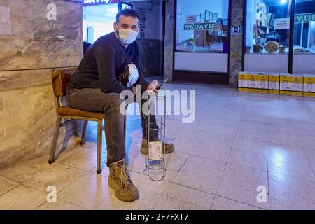 Giovane turco che indossa una maschera protettiva si siede su una sedia vicino al negozio con uno smartphone in mano durante l'infezione da coronavirus covid-19. Foto Stock