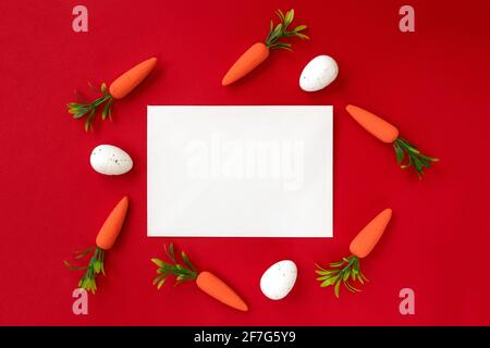 Layout creativo fatto di uova di Pasqua e carote con carta bianca su sfondo rosso. Sfondo pasquale minimo. Concetto di vacanze di primavera. Foto Stock