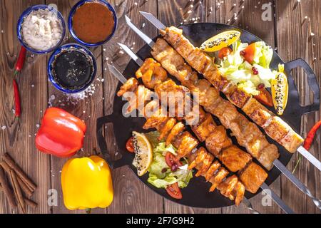 Kebab tritato su uno spiedino di metallo con salsa, cetrioli tagliati a fette, pomodori e cipolle su un vassoio di legno Foto Stock