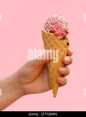 Tenere a mano il gelato alla fragola nel cono del waffle con gli spruzzi di caramelle isolati su sfondo rosa chiaro, il gusto del concetto estivo, dessert congelato Foto Stock