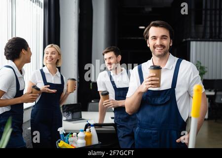 Detergente positivo che tiene la tazza di carta e guarda la fotocamera vicino colleghi interrazziali e detergenti in ufficio Foto Stock