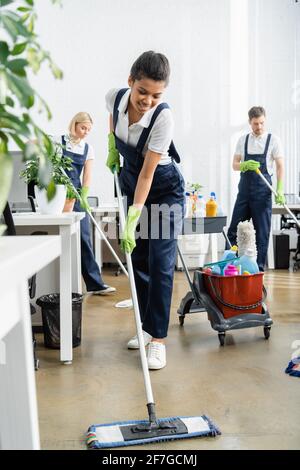 Positivo afroamericano lavaggio pavimento vicino pianta su sfocato in primo piano in ufficio Foto Stock