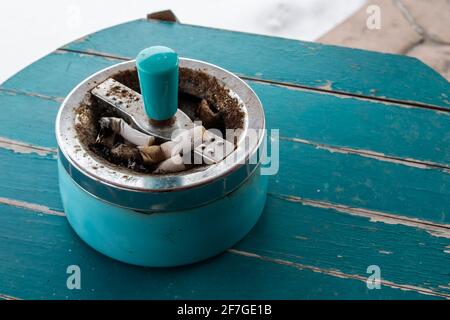 Un posacenere retrò, antico, pieno di mozzi su un tavolo di legno sabbiato turchese. Vista laterale angolare. Foto Stock