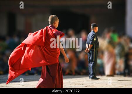 Regno del Bhutan, Asia, Himalaya. In attesa di gente gente folla in background. Di fronte a un poliziotto di guardia di sicurezza. Un giovane monaco si avvicina Foto Stock