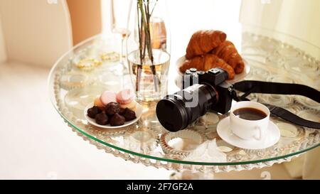 Fotocamera con un obiettivo su un tavolo di vetro. Tazza di caffè, macaron e croissant Foto Stock