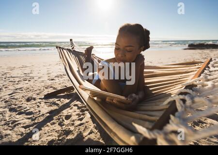 Donna afro-americana felice che si trova in amaca sulla spiaggia utilizzando tablet Foto Stock