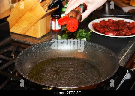 Primo piano di una mano che aggiunge olio ad una padella su un piano di cottura. Piatto di marinare manzo e alcune verdure verdi, così come altri articoli da cucina. Foto Stock