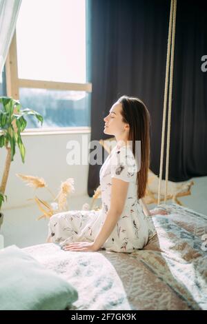 La giovane donna si siede sul letto in vestaglia e medita accanto alla finestra. Foto Stock