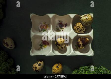 Uova di quaglia macchiate nel vassoio di uova floreali in porcellana bianca su sfondo verde scuro. Creatività e fotografia RAW. Foto Stock