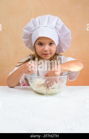 Una bambina vestita come cuoco impasta l'impasto. Cucina bambino stile di vita concetto. Il capretto ama, ha divertimento, studia e gioca nella cucina Foto Stock