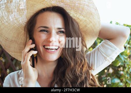 felice giovane donna in camicia bianca con cappello che parla su uno smartphone in terrazza. Foto Stock
