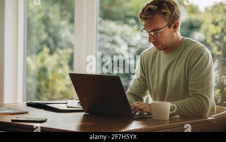Giovane uomo che lavora su un computer portatile. Uomo seduto al tavolo e che usa il computer portatile mentre sei a casa. Foto Stock