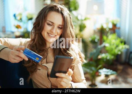 Green Home. Felice trendy donna di mezza età con lunghi capelli ondulati con carta di credito facendo shopping online sul sito di e-commerce nella casa moderna in su Foto Stock