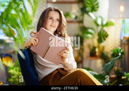 Green Home. Pendive alla moda casalinga di 40 anni con lunghi capelli ondulati con libro in pantaloni verdi e blusa grigia nella casa moderna in giornata di sole. Foto Stock