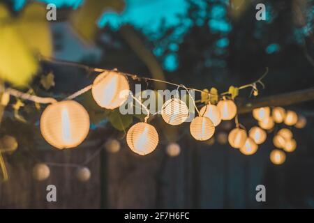 Fila di lanterne rotonde di carta in un giardino di casa su uno sfondo di legno paglia di notte. Decorazioni festive in giardino una sera. Foto Stock