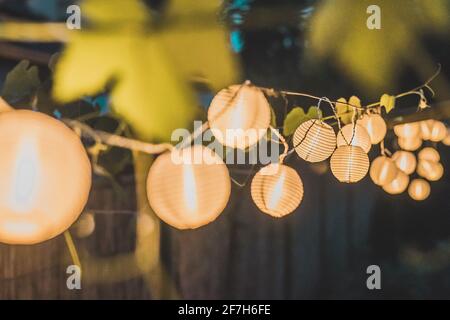 Fila di lanterne rotonde di carta in un giardino di casa su uno sfondo di legno paglia di notte. Decorazioni festive in giardino una sera. Foto Stock