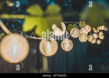 Fila di lanterne rotonde di carta in un giardino di casa su uno sfondo di legno paglia di notte. Decorazioni festive in giardino una sera. Foto Stock