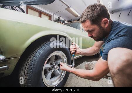 Giovane uomo che rimuove ruote o pneumatici da una vecchia auto d'epoca degli anni '60 o '70 nel suo garage di casa. Gli strumenti sono visibili in giro. Foto Stock