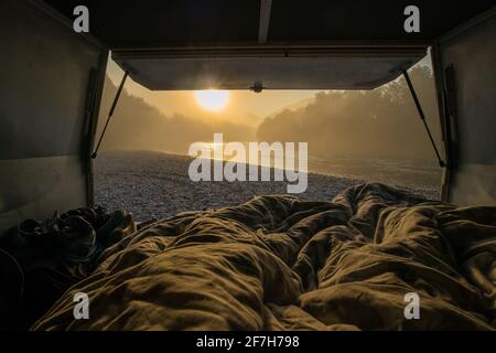 Vista da un letto nel campervan d'epoca parcheggiato sulle rive di un fiume nelle prime ore del mattino. Pernottamento romantico in un campeggio selvaggio vicino ad un r Foto Stock